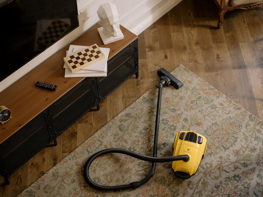 A person using a yellow vacuum cleaner with a black hose to perform surface cleaning and deep cleaning of an ornate, patterned area rug in a living room. The rug features intricate floral designs in m