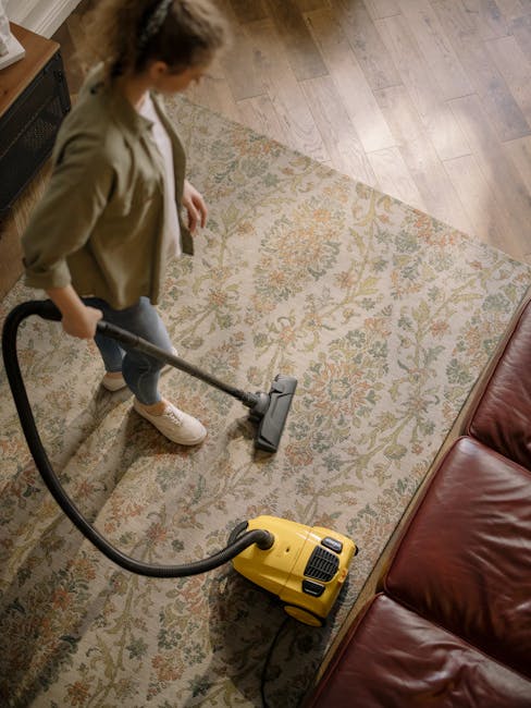 A woman with light brown hair tied back is vacuuming a patterned area rug situated on a wooden floor in a living room with natural light. She is wearing a beige jacket, white top, grey pants, and whit