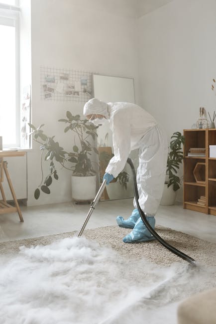 A woman with light brown hair tied back is vacuuming a patterned area rug situated on a wooden floor in a living room with natural light. She is wearing a beige jacket, white top, grey pants, and whit