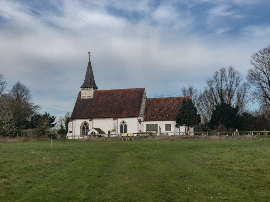 Photograph of St Marys Church Cranham, a traditional white-painted church with a tall, steeply-pitched wooden spire topped with a weather vane. The church features pointed arch stained glass windows a